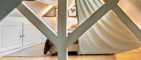 A loft bedroom with a sloped ceiling, a wooden floor, and a bed. There is a ladder against the wall with a teddy bear on it.