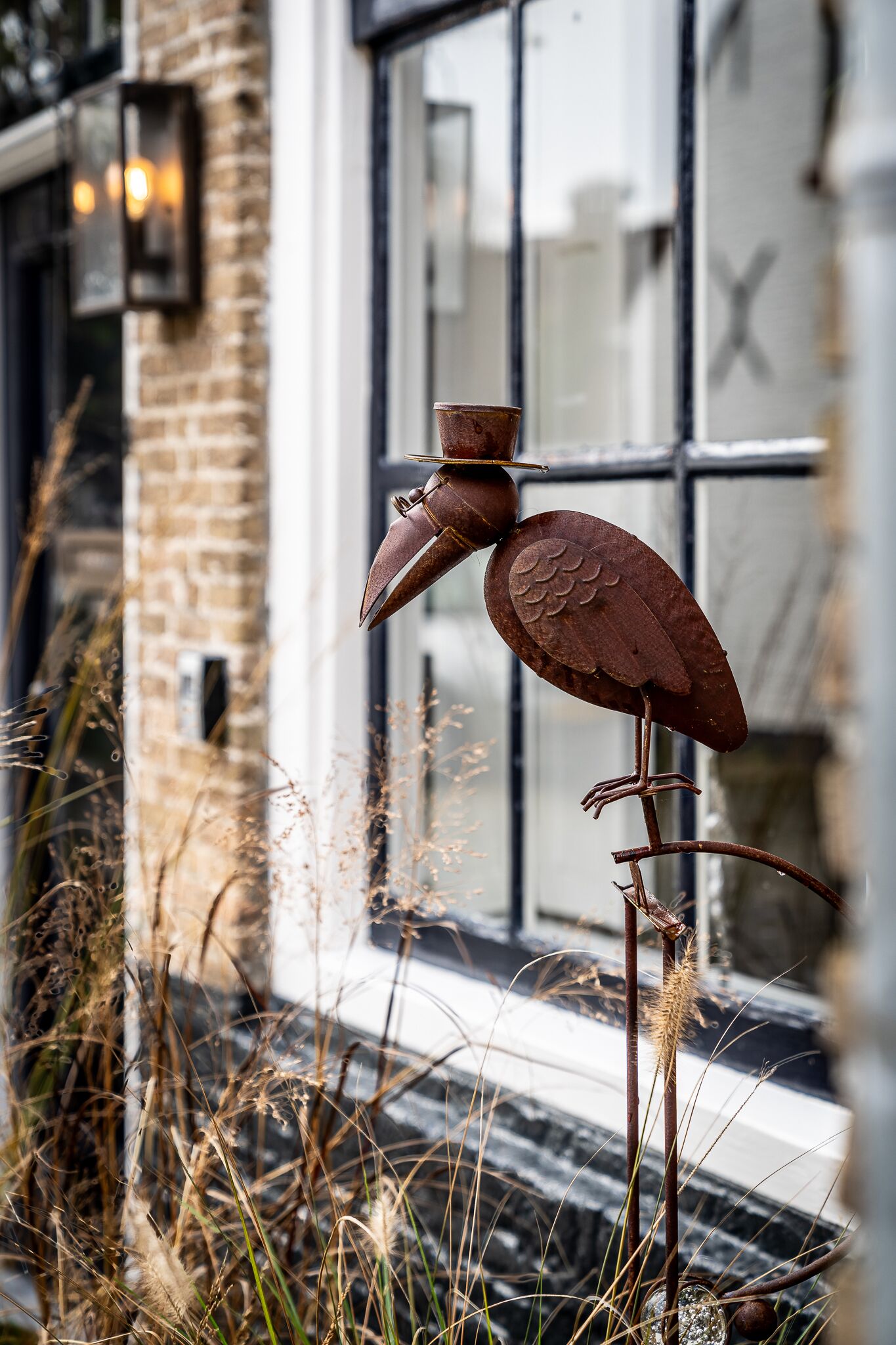 A metal bird sculpture with a large beak and wings, positioned on a metal rod in front of a building with a brick facade and large windows.