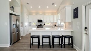 A modern kitchen with white cabinetry, a stainless steel refrigerator, a central island with bar stools, and recessed ceiling lighting. - Sea Bliss - 3 bedroom, 2 bath home that sleeps 8 - Easy Beach Access! (Crystal Beach)