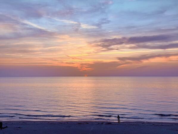Eine Strandansicht mit Sonnenuntergang über dem Meer, eine Person steht am Ufer, und eine ferne Sonne geht am Horizont unter.
