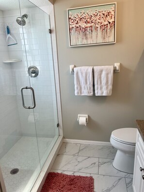 Ein Badezimmer mit Duschkabine aus Glas, Toilette, Marmorboden, rotem Teppich und einem Handtuchhalter mit zwei Handtüchern.