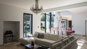 A spacious living room with a large beige sofa, a coffee table, and a chandelier. There is a dining area with a red table and chairs visible through large windows. - Beautiful contemporary villa between lake and ocean. Free wifi. (Soorts-Hossegor)