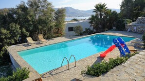 A swimming pool with a red slide, surrounded by stone walls and greenery, with a view of a mountain and water in the background. - Blue Paradise: Comfy Beach Villa walking distance to the sea / pool access (Paros)