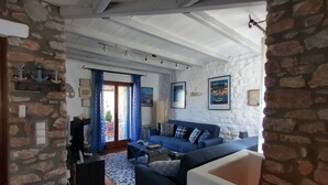 A living room with a stone wall, a blue sofa, a coffee table, and a view through a glass door. - Blue Paradise: Comfy Beach Villa walking distance to the sea / pool access (Paros)