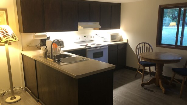 Una cocina compacta con armarios oscuros, microondas, tostadora y fregadero. Hay un comedor con mesa y sillas.