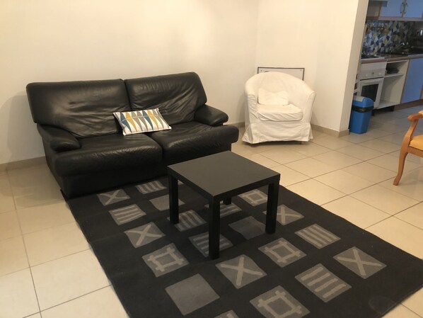 A living room with a black leather sofa, a small black table, and a white armchair. A patterned rug is on the floor.