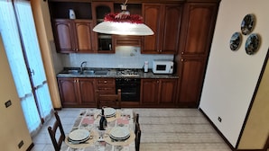 A dining area with a table set for four, a kitchen in the background, and a ceiling light fixture. - Villetta Camelia with private parking just a stone's throw from Lucca (Capannori)