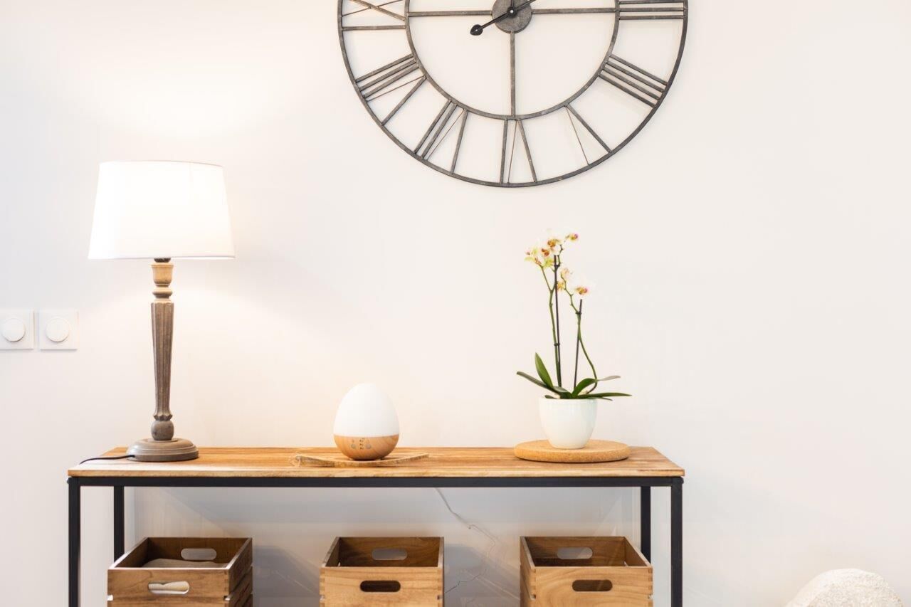 Una mensola in legno con un orologio a parete con numeri romani, una lampada, un uovo decorativo e una pianta in vaso.