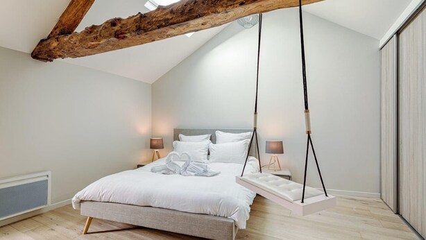 A bedroom with a sloped ceiling, a bed with white bedding, two bedside lamps, and a hanging swing chair.