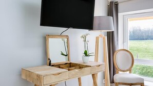 A room with a wooden desk, a television mounted above it, a chair, and a window with a view of a green field.