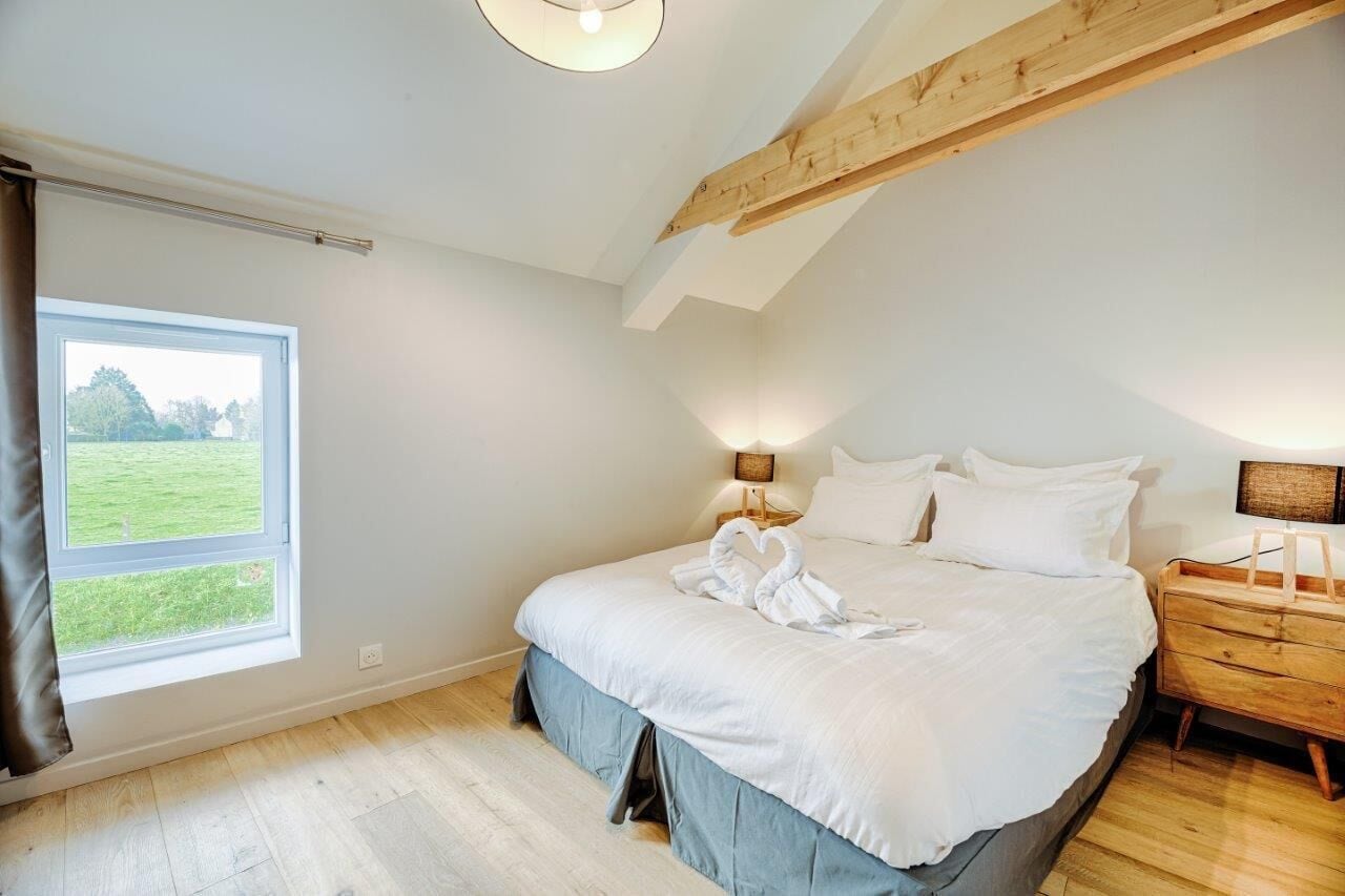 Ein Schlafzimmer mit einem großen Bett, einem Holzbalken an der Decke, einem Fenster mit Blick auf ein grünes Feld und einem Nachttisch mit einer Lampe.