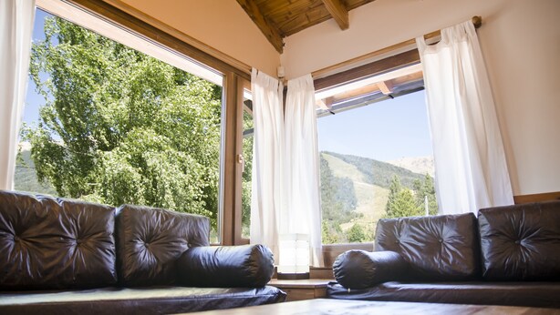 Ein Zimmer mit Ledersofa, das einer großen, mit Vorhängen versehenen Fenster gegenüberliegt, einem Holztisch im Vordergrund und Blick auf grüne Hügel.