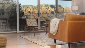 A modern living room with a leather sofa, a woven chair, and a large window offering a view of a mountainous landscape. - Sunrise to moonrise views. Hike/bike out the door. (Sedona)