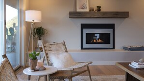 A cozy living room with a fireplace, a comfortable chair, a side table with a lamp, and a view through a large window. - Sunrise to moonrise views. Hike/bike out the door. (Sedona)