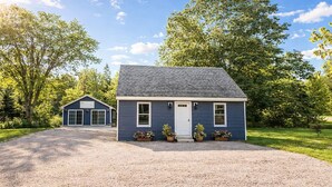 A two-story house with a gravel driveway and a front porch with potted plants. - Gateway to Acadia Renovated Milbridge Coastal Home (Milbridge)