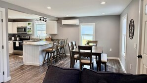 A modern kitchen with white cabinets, a central island, and a dining area with wooden chairs. - Gateway to Acadia Renovated Milbridge Coastal Home (Milbridge)
