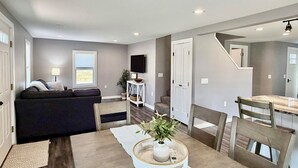 A modern living room with a sofa, dining table, chairs, a TV, and a staircase. - Gateway to Acadia Renovated Milbridge Coastal Home (Milbridge)
