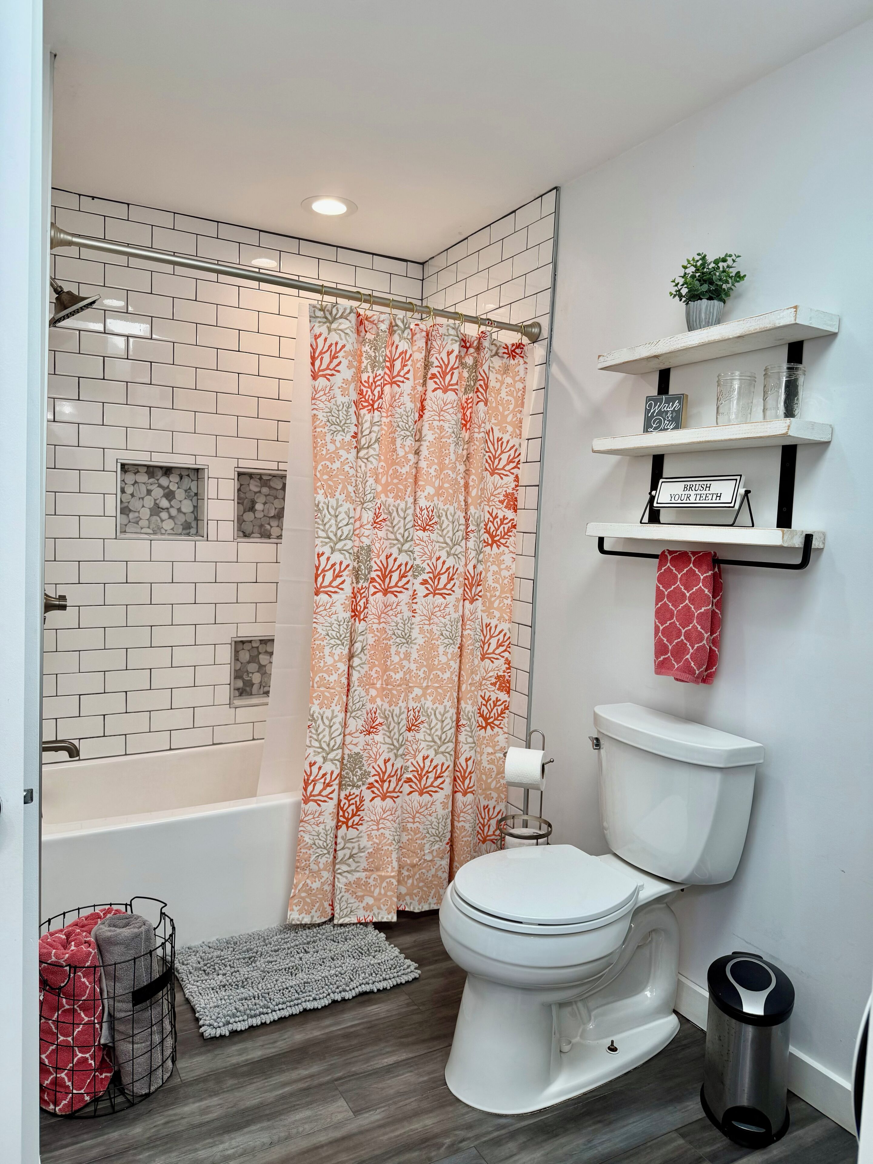 Un baño con cortina de ducha, inodoro, estante con frascos y una planta pequeña.