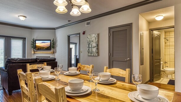 Uma sala de jantar com uma mesa de madeira posta para seis pessoas, rodeada por cadeiras de madeira. Há um sofá de couro preto ao fundo, uma televisão fixada na parede e um quadro sobre a porta.
