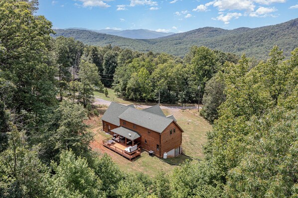 A wooden cabin with a deck and outdoor seating, surrounded by dense forest and mountains in the background.