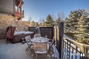 Relax in the patio area with a hot tub, table, and chairs, surrounded by a wooden fence and evergreen trees.
