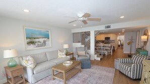 A living room with a sofa, coffee table, and ottoman, a striped armchair, a painting, and a staircase leading to a kitchen. - Luxury 3BD Beachfront Living: La Playa 203D by RVA (Longboat Key)