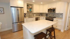 A modern kitchen with white cabinetry, a stainless steel refrigerator, a microwave, and a central island with stools. - Luxury 3BD Beachfront Living: La Playa 203D by RVA (Longboat Key)