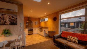 A modern kitchen with wooden cabinets, a dining area with white chairs, and a cozy living room with a leather sofa and a large window.