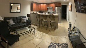 A compact living space with a sofa, a glass coffee table, a kitchen area with a refrigerator and bar stools, and a TV mounted on the wall. - JC's Place #1 (Hebron)
