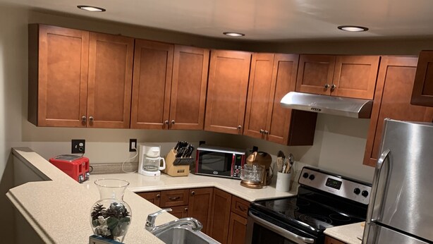 A modern kitchen with wooden cabinets, a stainless steel refrigerator, a built-in oven, and a sink. There is a welcome sign on the counter and a bar stool area. - JC's Place #1 (Hebron)