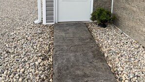 A house entrance with a concrete pathway leading to a white door, surrounded by gravel and a small potted plant. - JC's Place #1 (Hebron)