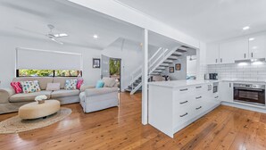 Amplia sala de estar con sofá, reposapiés y cocina con muebles blancos y horno de acero inoxidable.