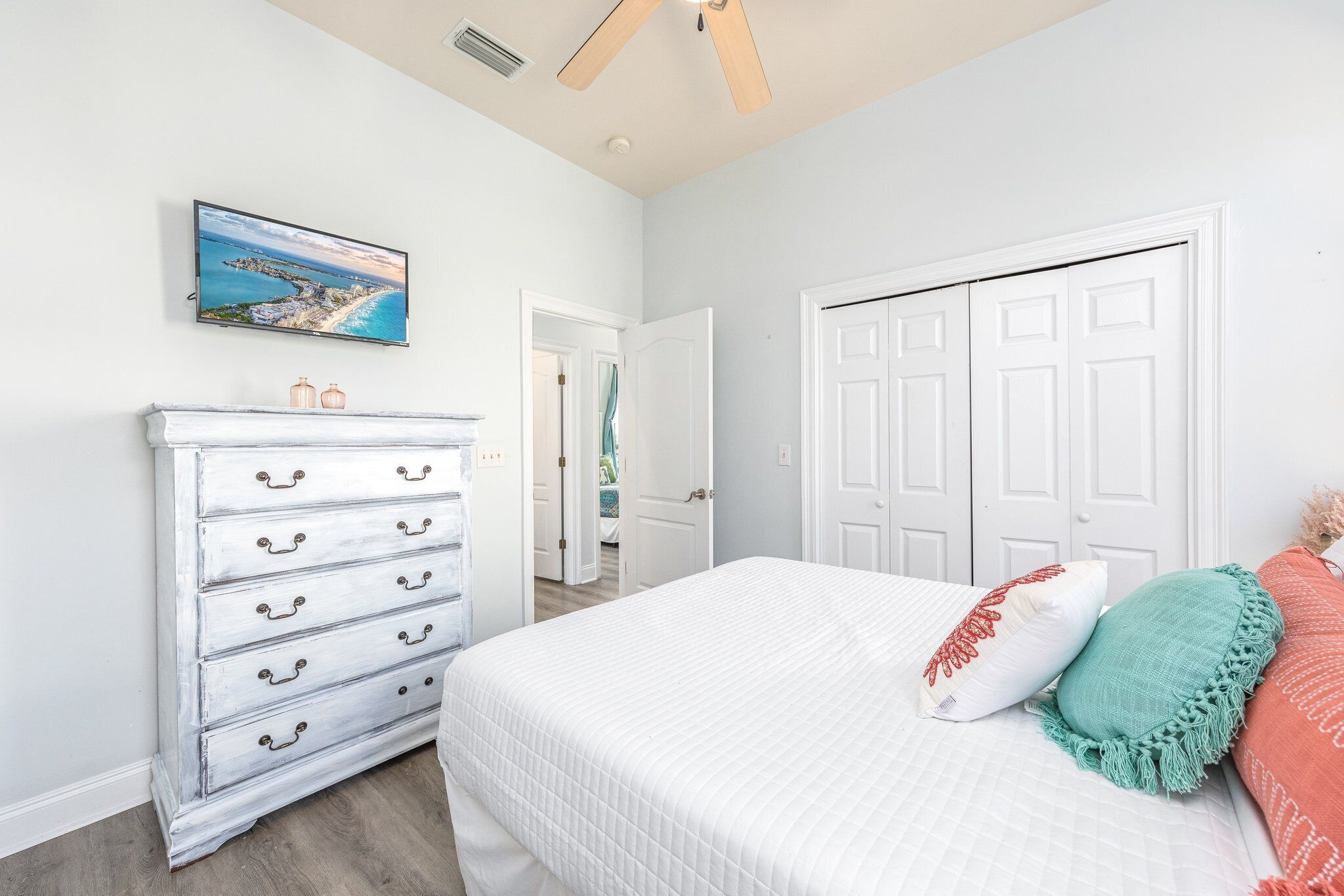 A bedroom with a bed, a dresser, a ceiling fan, a painting, and a door.