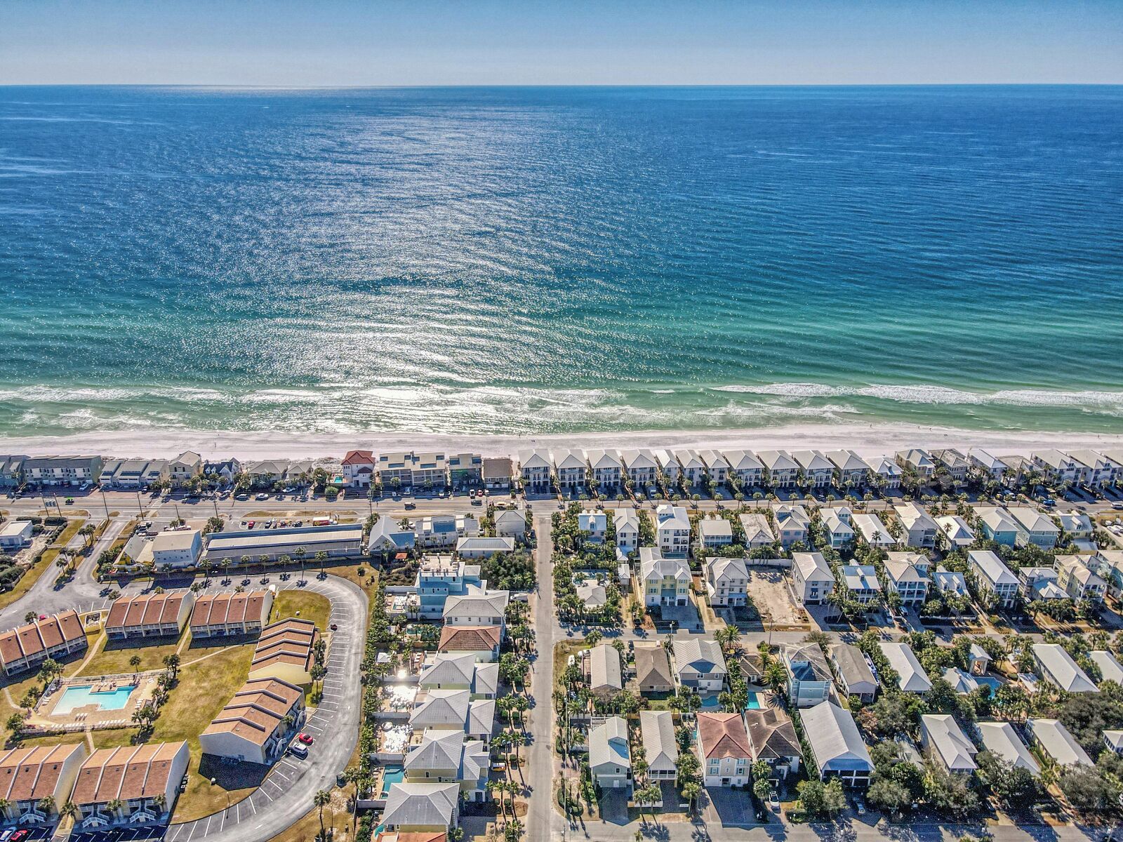 An aerial view of a coastal residential area with beachfront properties, a swimming pool, and a road leading to the beach.