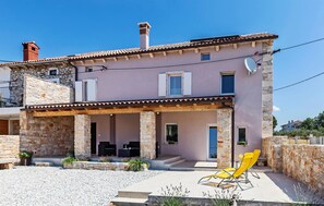 Una casa de dos plantas con fachada de piedra, techo de tejas y un porche cubierto con mobiliario exterior.