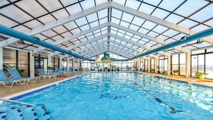Une grande piscine intérieure dotée d’un toit en verre, de chaises longues bleues et de plantes en pot.