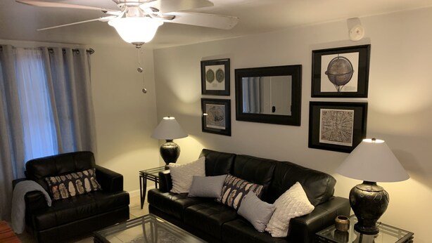 A living room with a black leather sofa, two side tables with lamps, a glass coffee table, a ceiling fan, and framed artwork on the walls. - JC's Place #2 (Hebron)