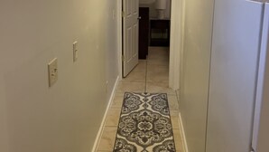A narrow hallway with a patterned rug, a ceiling light, and a door leading to a room with a lamp and a cabinet. - JC's Place #2 (Hebron)