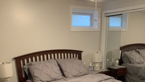 A bedroom with a wooden bed, a nightstand with a lamp, a bedside table with a lamp, and a window with blinds. - JC's Place #2 (Hebron)