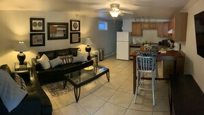 A living room with a sofa, coffee table, and a dining area with a chair and table. There is a kitchen with cabinets and appliances. - JC's Place #2 (Hebron)