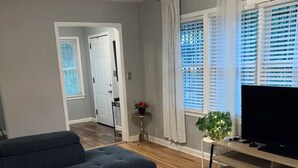 Una sala de estar moderna con un sofá oscuro, una mesa de centro de vidrio con flores, un televisor sobre un soporte de madera y un ventilador de techo.