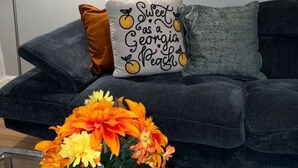 Una sala de estar moderna con un sofá gris, un cojín decorativo, un jarrón con flores anaranjadas y un cuadro en la pared.