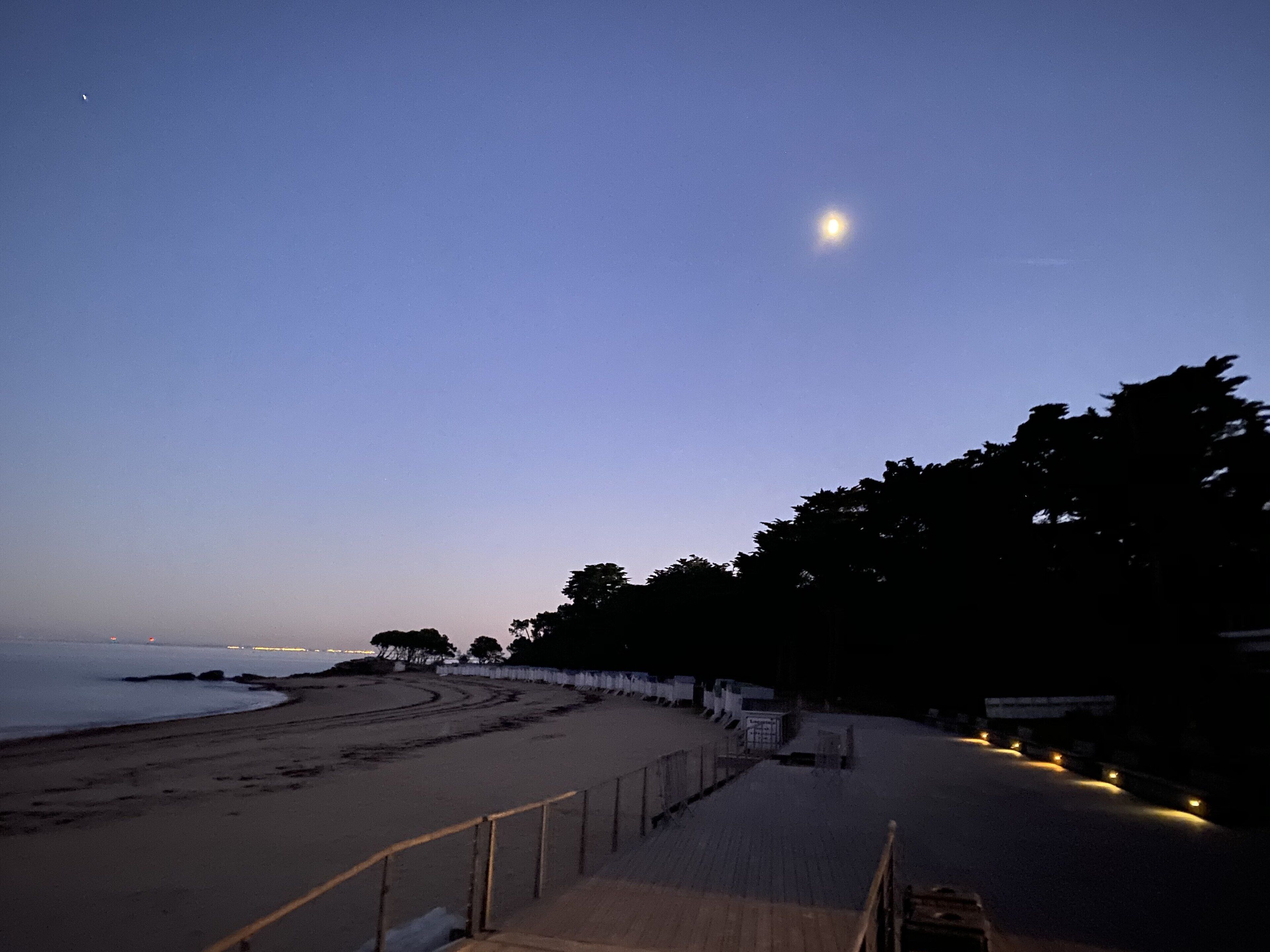 Una playa con un camino de madera que lleva al agua, una cerca a un lado y una luna en el cielo.