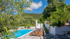 A swimming pool with lounge chairs, surrounded by greenery and a view of hills.