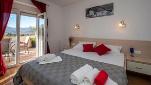 A hotel room with a bed, two bedside tables, a window with red curtains, a view of the outdoors, and a painting on the wall.