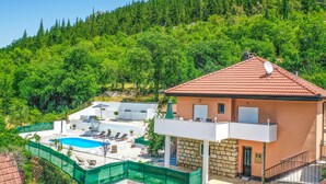 A two-story house with a swimming pool, surrounded by lush greenery and a forested hillside.