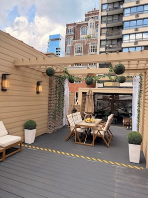 Una terraza en la azotea con una mesa y sillas de madera, plantas en macetas y vistas del paisaje urbano.