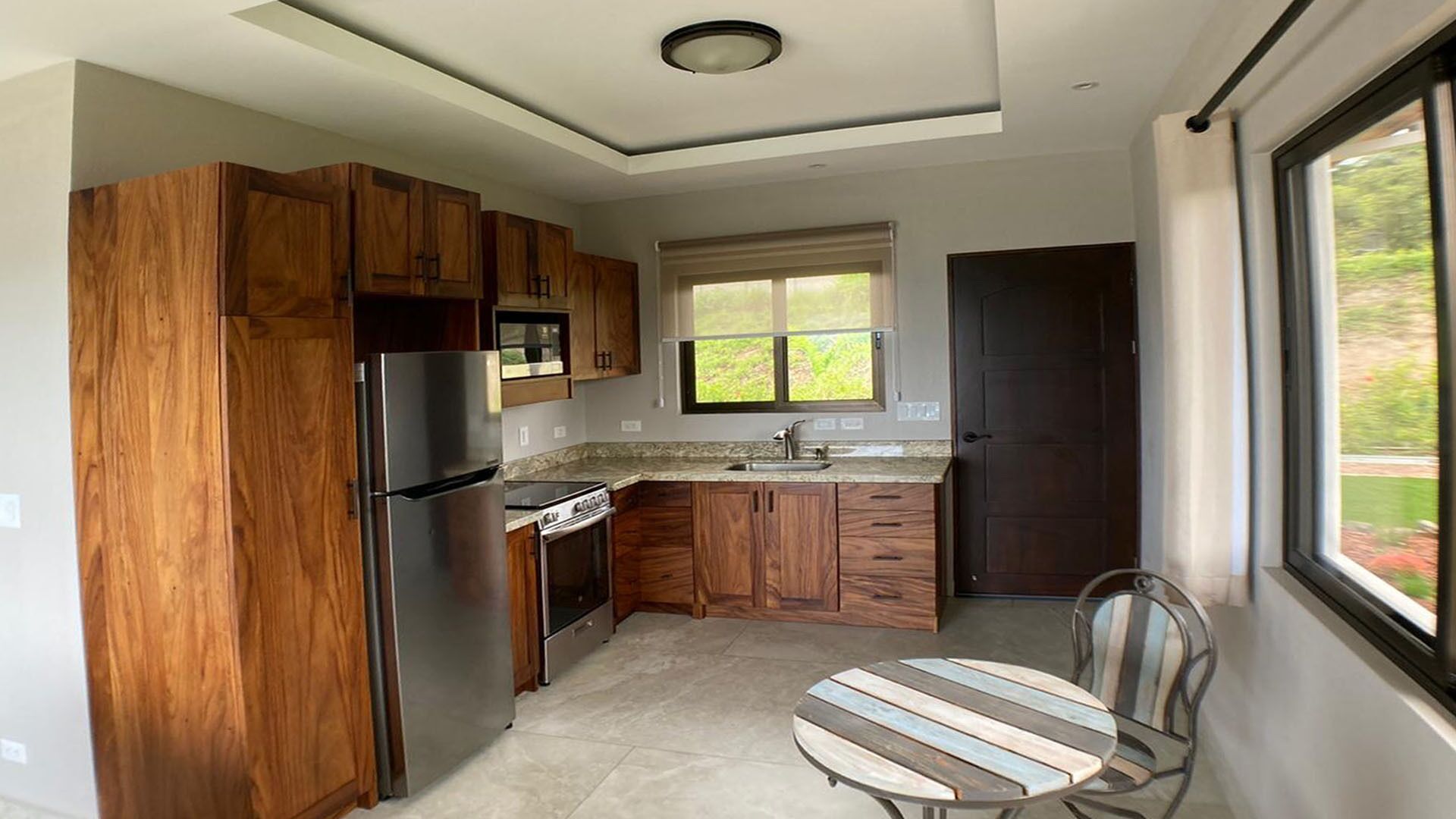 A compact kitchen with wooden cabinets, a stainless steel refrigerator, a stove, and a window with a view of greenery.