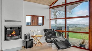 A modern living room with a fireplace, a comfortable lounge chair, and a large window offering a view of the outdoors. - Nice home in Sjølund with sauna (Sjølund)