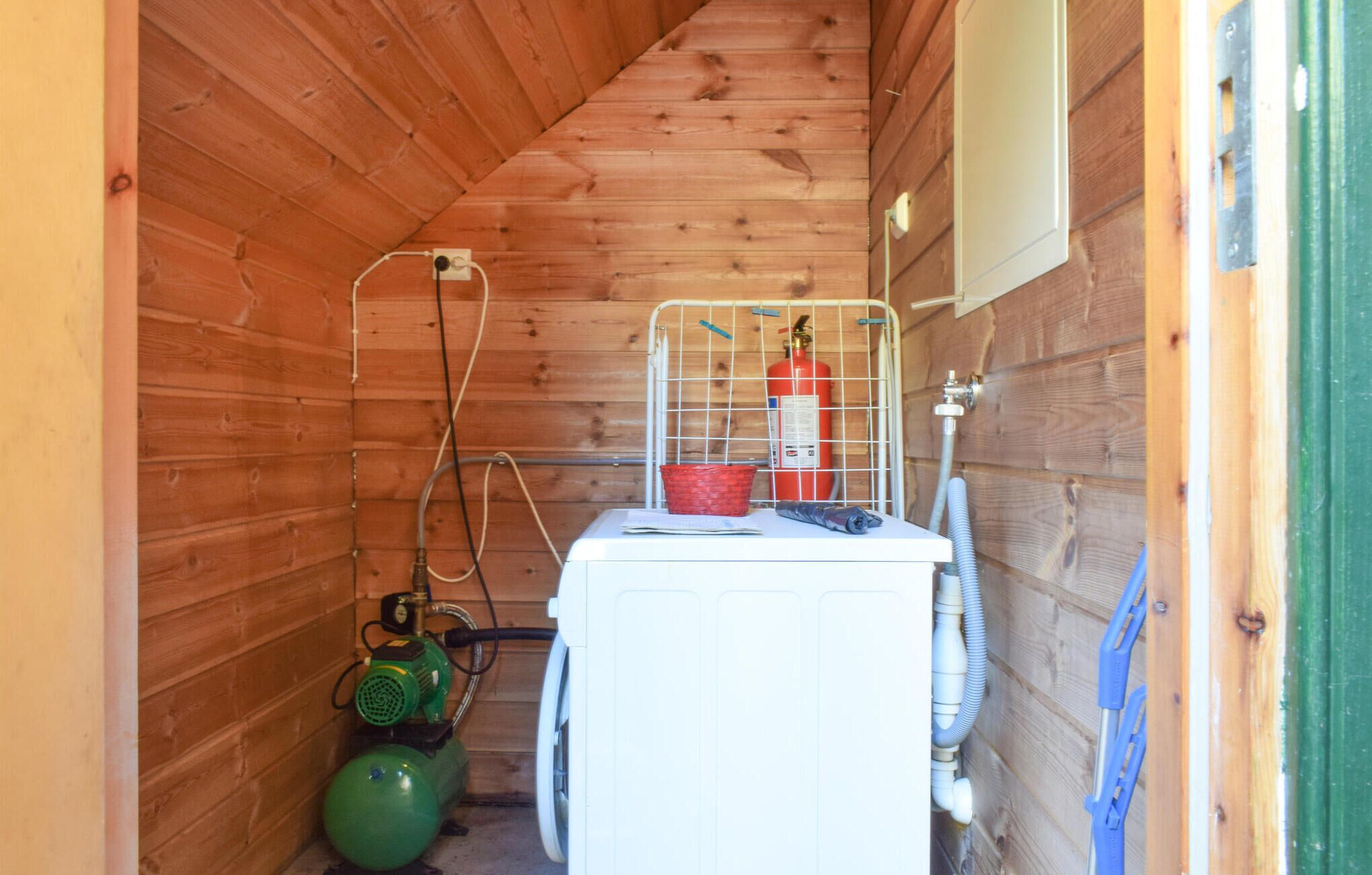 Une petite pièce fermée, servant de local technique, avec des murs en bois, un lave-linge blanc, une extincteur rouge et une bouteille de gaz verte.
